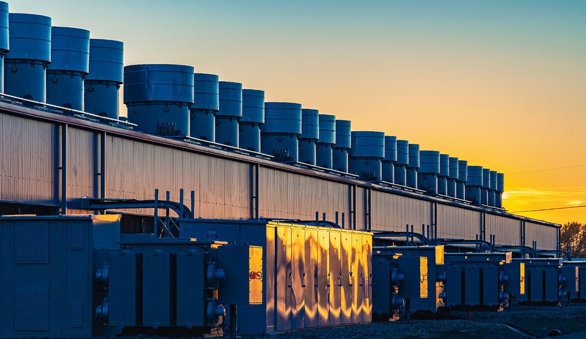 The shiny exterior of an industrial building stands against an orange sky. The sky is reflected in the building’s exterior.