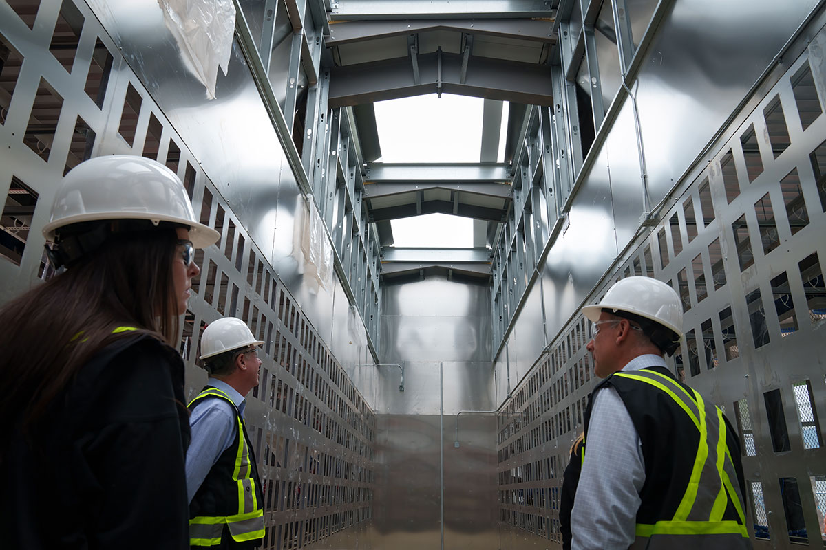Three people wearing white hardhats stand in a towering room. The interior is clad in stainless steel.