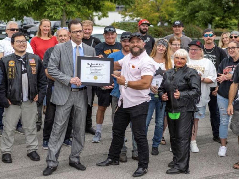 Two men pose with a framed honorary degree. They are outside, and surrounded by a group of bikers.