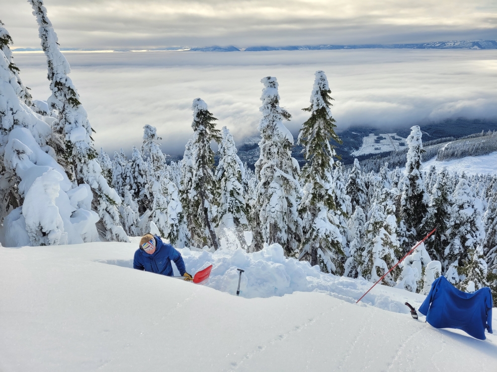 In the foreground, a person in a ski jacket stands waist-deep in a snow pit, using a red shovel to remove snow. Avalanche gear, like a saw and probe, sit nearby. In the background, the treed slope drops steeply away toward toward a mountain valley shrouded in cloud.
