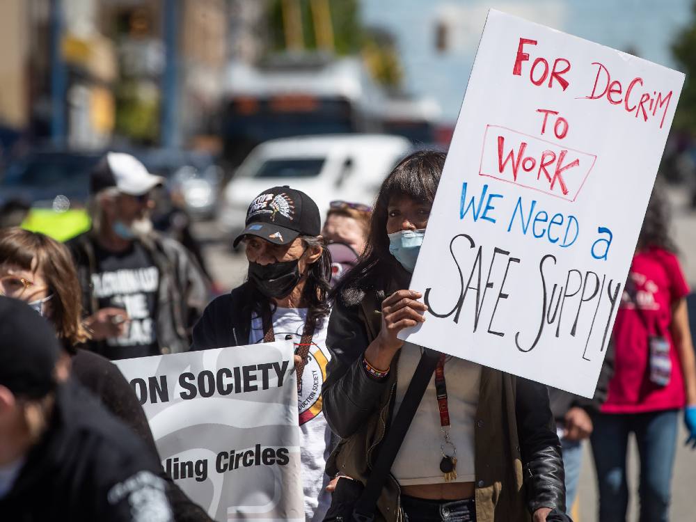 A woman with dark hair holds a white rectangular poster board that reads &ldquo;For decrim to work we need a safe supply.&rdquo; She is marching in a protest among others holding a white whose black text is partially visble in the photo. It&rsquo;s a bright, sunny day.