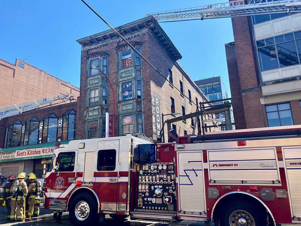 A fire truck is parked in front of a brick building called the Keefer Rooms. Firefighters wearing yellow firefighting uniforms gather near the front of the truck.