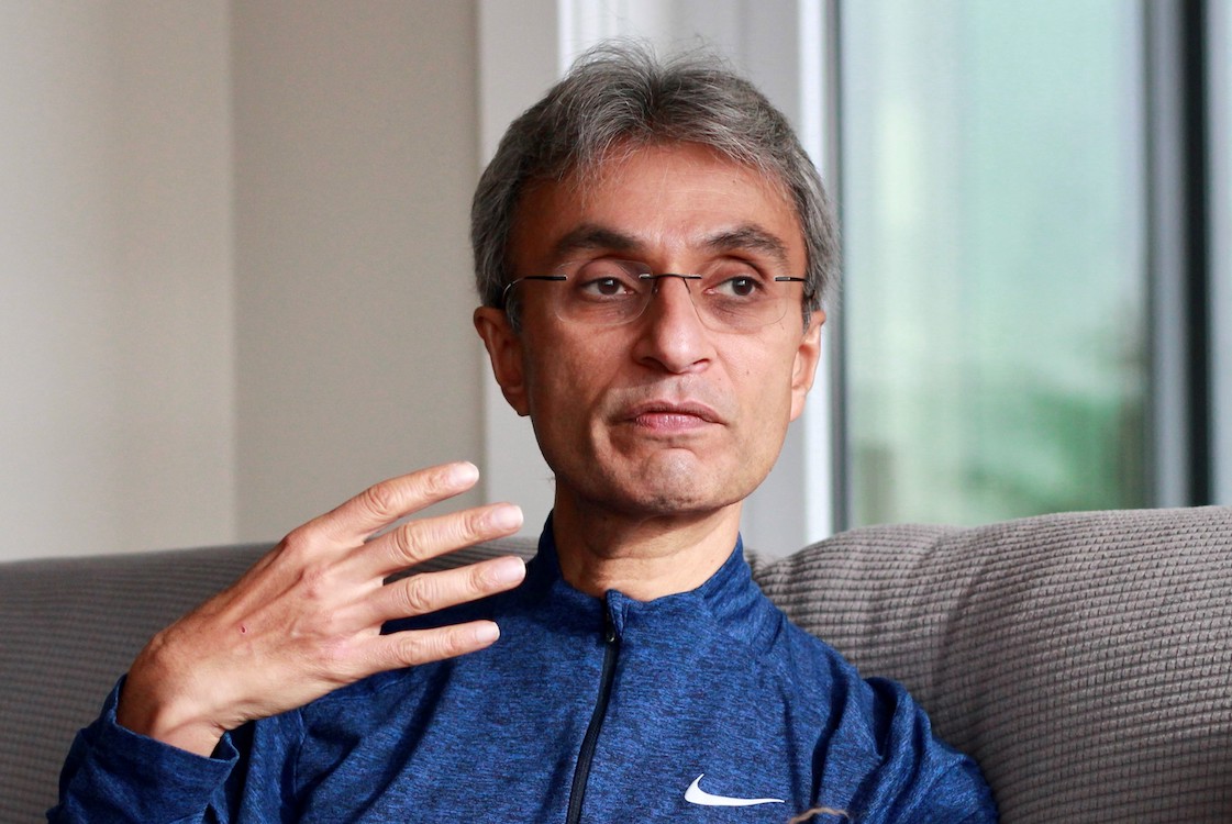 A man sits on a grey couch. He's using his hands as he speaks. 