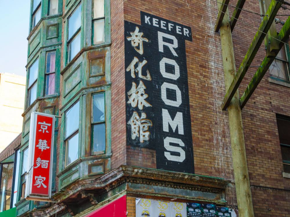 The side of an old building in Vancouver&rsquo;s Chinatown. There is a teal-painted fa&ccedil;ade with bay windows at the front. The side is brick and has &ldquo;Keefer Rooms&rdquo; painted on it in white in both English and Chinese.