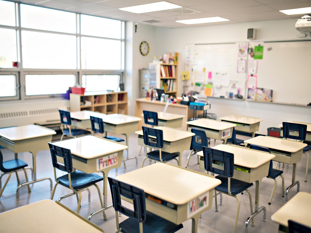 An empty elementary school classroom, full of desks and blue chairs. Colourful resources are taped to the whiteboard at the front of the room.