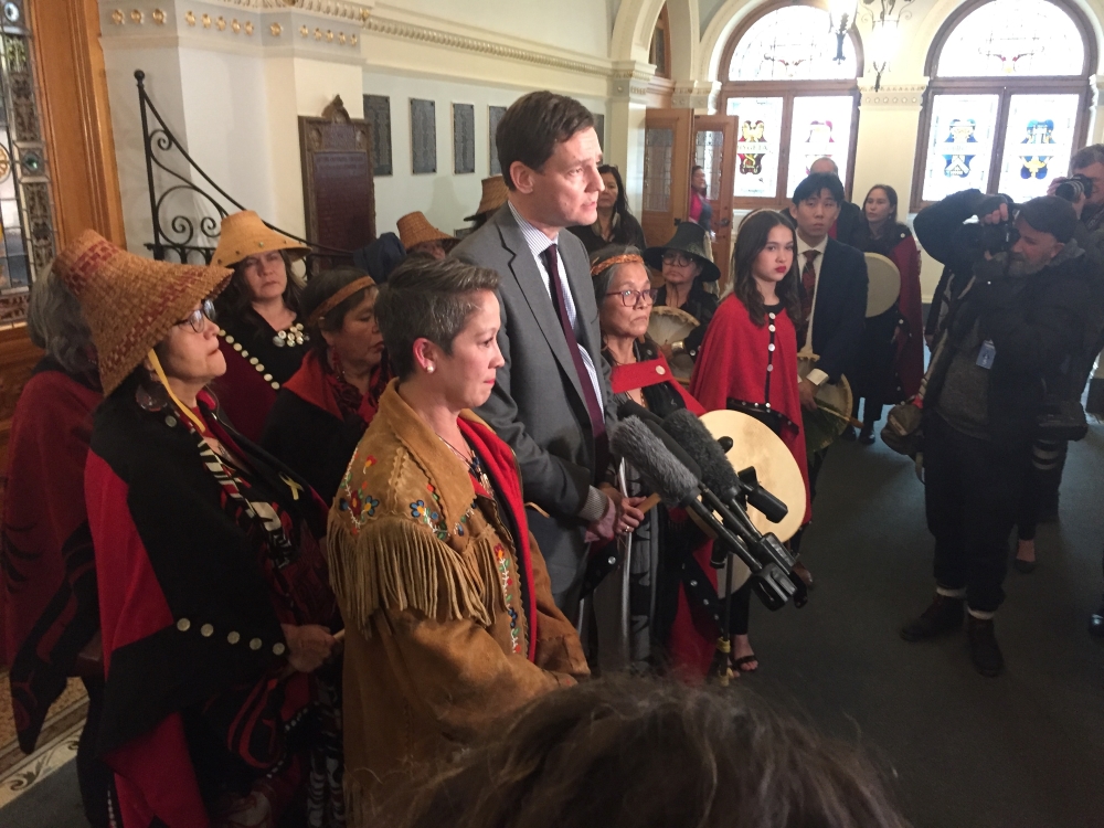 A woman with short hair and a beaded, fringed jacket stands beside a tall man in a grey suit, white shirt and red tie. Behind them are women wearing cedar hats and other traditional regalia and in front media photographers.
