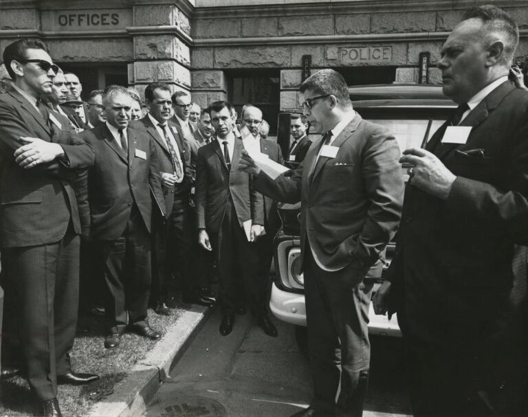 A black-and-white photo shows a man with a crewcut and glasses reading from a piece of paper, as other men in suits, white shirts and ties listen. In the background is a stone building.