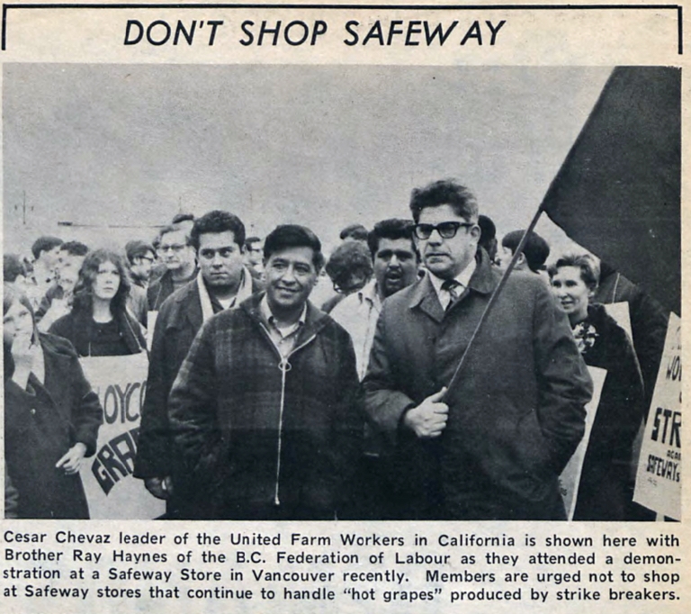 A black-and-white photo shows a group of people. In the front is Cesar Chavez, wearing a paid jacket, and Ray Haynes, wearing a dark trench coat and shirt and tie and holding a flag.