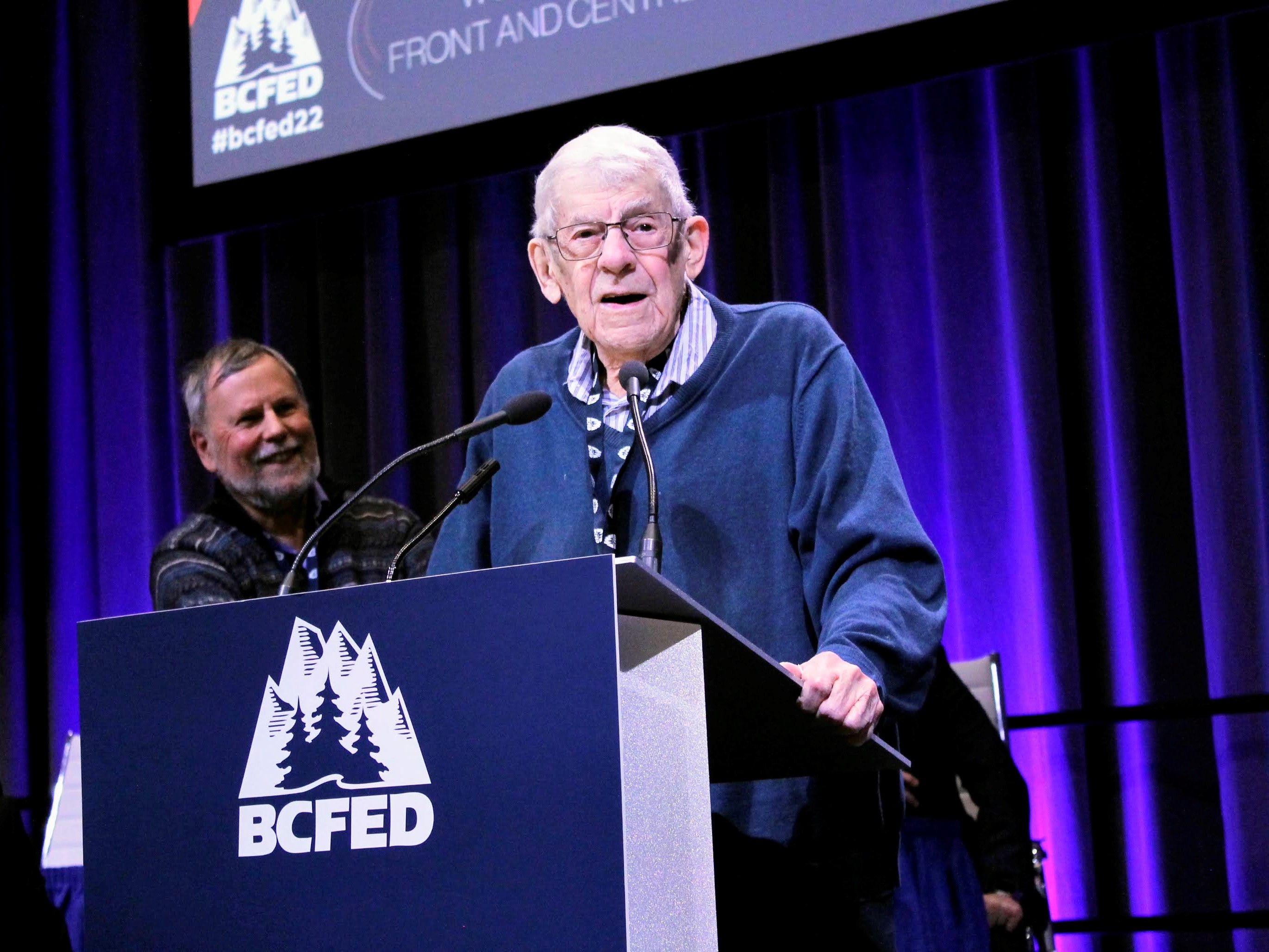 An elderly man wearing a navy blue sweater stands at a podium reading ‘BCFED.’ A royal blue curtain is behind him.