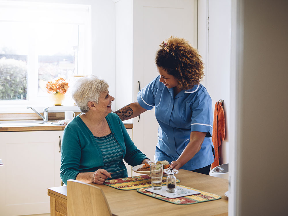 An elderly woman sits in a sunlit room in front a kitchen table. A care nurse serves her food.