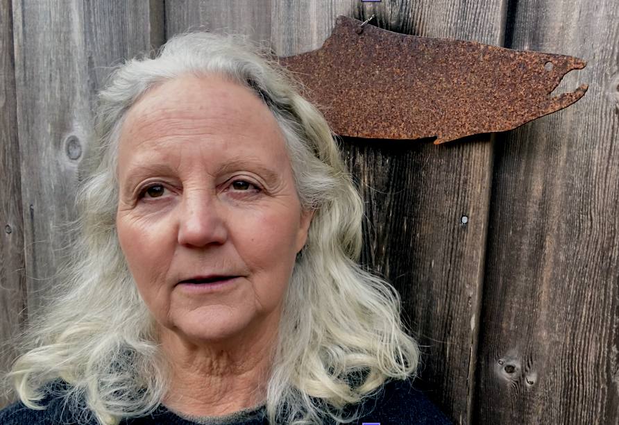 Alexandra Morton, a white woman in her senior years, with long white hair, speaks to the camera against a wood wall on which hangs a rusted metal ornament in the shape of a salmon. 
