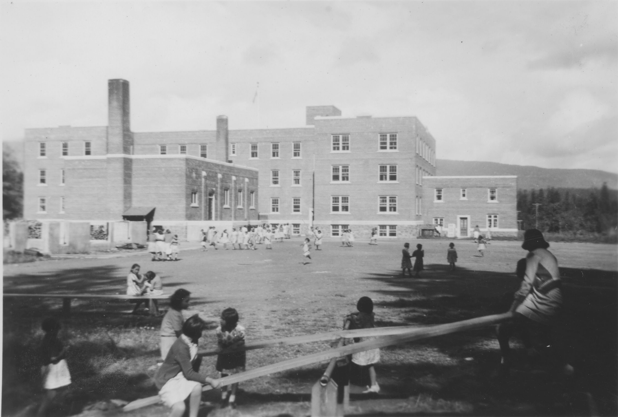 A black-and-white photo shows children on a teeter-totter in the foreground, with a large brick building in the background.