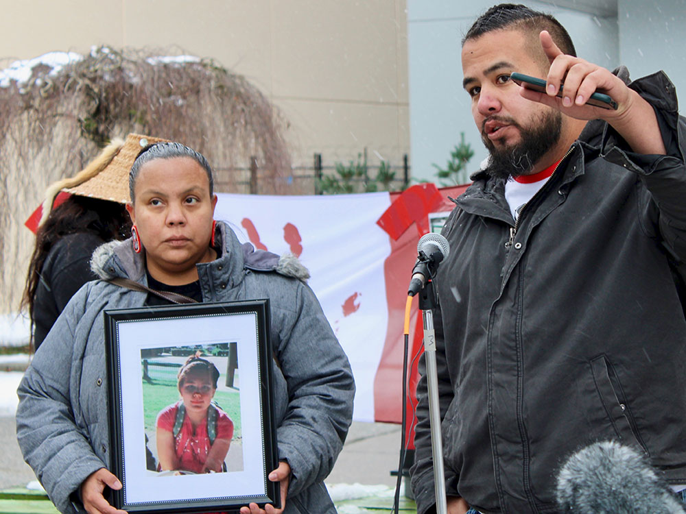 A serious looking person in a grey parka hold a photo of a young person. Beside her, a man wearing a black jacket holds a cell phone and points into the distance.