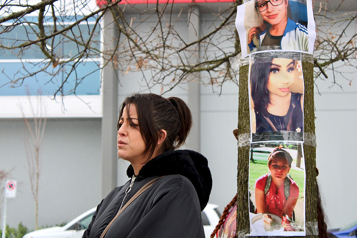A woman with black hair and wearing a black park looks into the distance with a sombre expression.