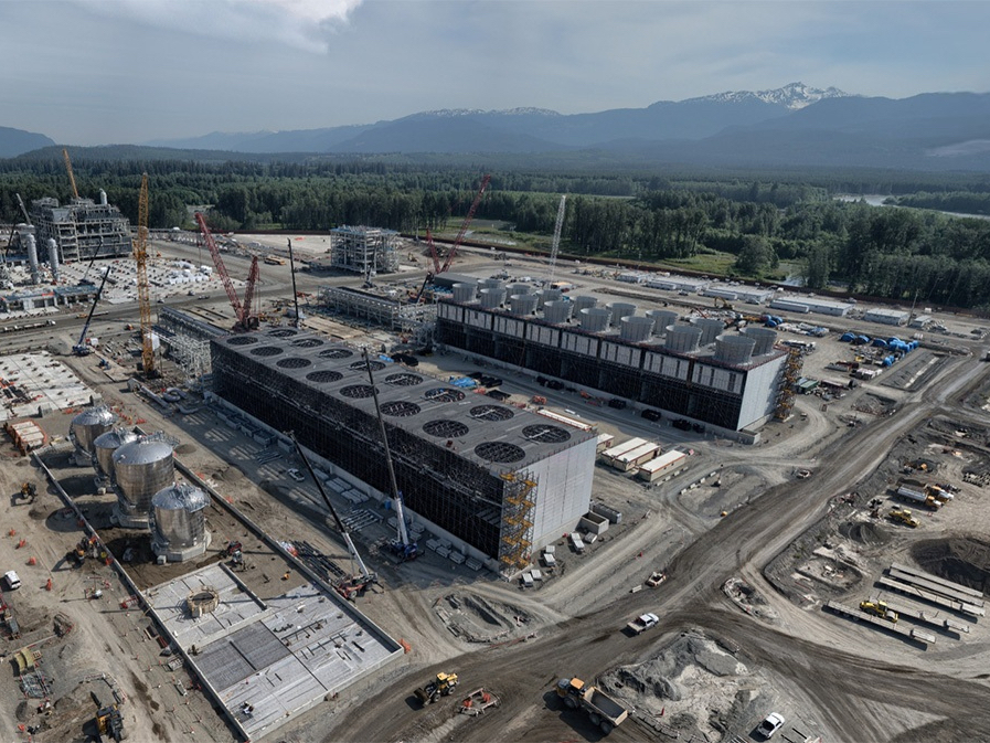 An aerial view shows the LNG Canada plant with large tanks and buildings on a large gravel work site.