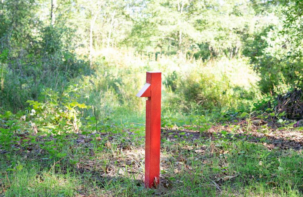 A short red structure stands in a sunny, grassy grove. It is a well that has been installed in the Kwikwetlem cemetery. It stands in the centre of the frame in a grassy patch, surrounded by green bushes. It&rsquo;s a sunny day. 