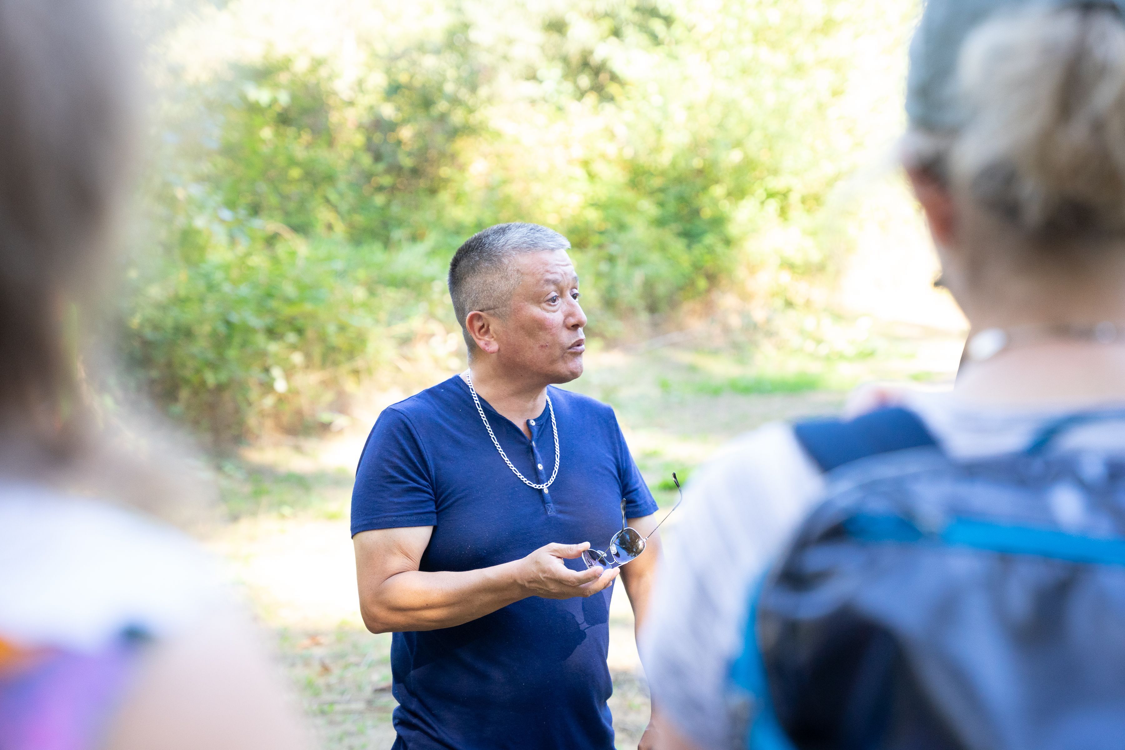 George Chaffee stands looking towards the right of the frame, mid-speech. He is holding a pair of sunglasses in his left hand and is wearing a blue T-shirt. Two people stand in soft focus in the foreground, part of a group&nbsp;he is leading. They are standing in a sunny green grove.
