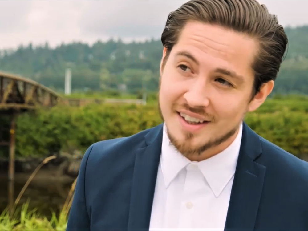 A young man in a crisp white shirt and blue suit jacket gestures on a river bank with green foliage. 