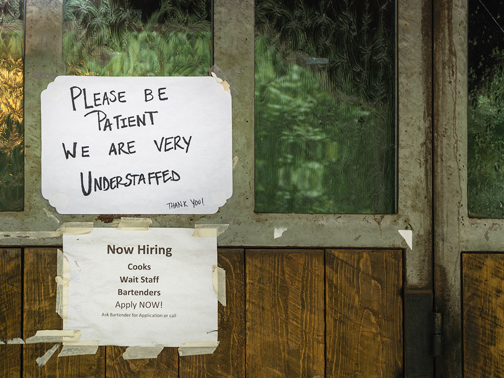 Two hand-lettered signs on a wooden bar door say &lsquo;Please Be Patient &mdash; We Are Very Understaffed&rdquo; and list a list of job vacancies.