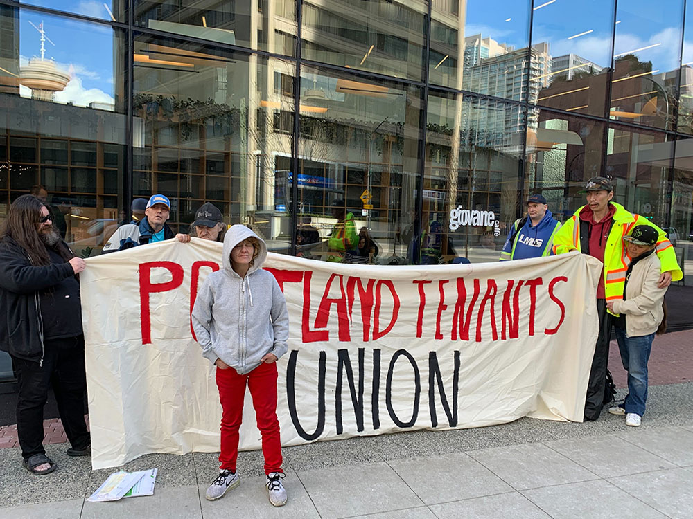 Eight people stand in front of a gleaming office tower holding a large cloth banner that reads &ldquo;Portland Tenants Union.&rdquo;
