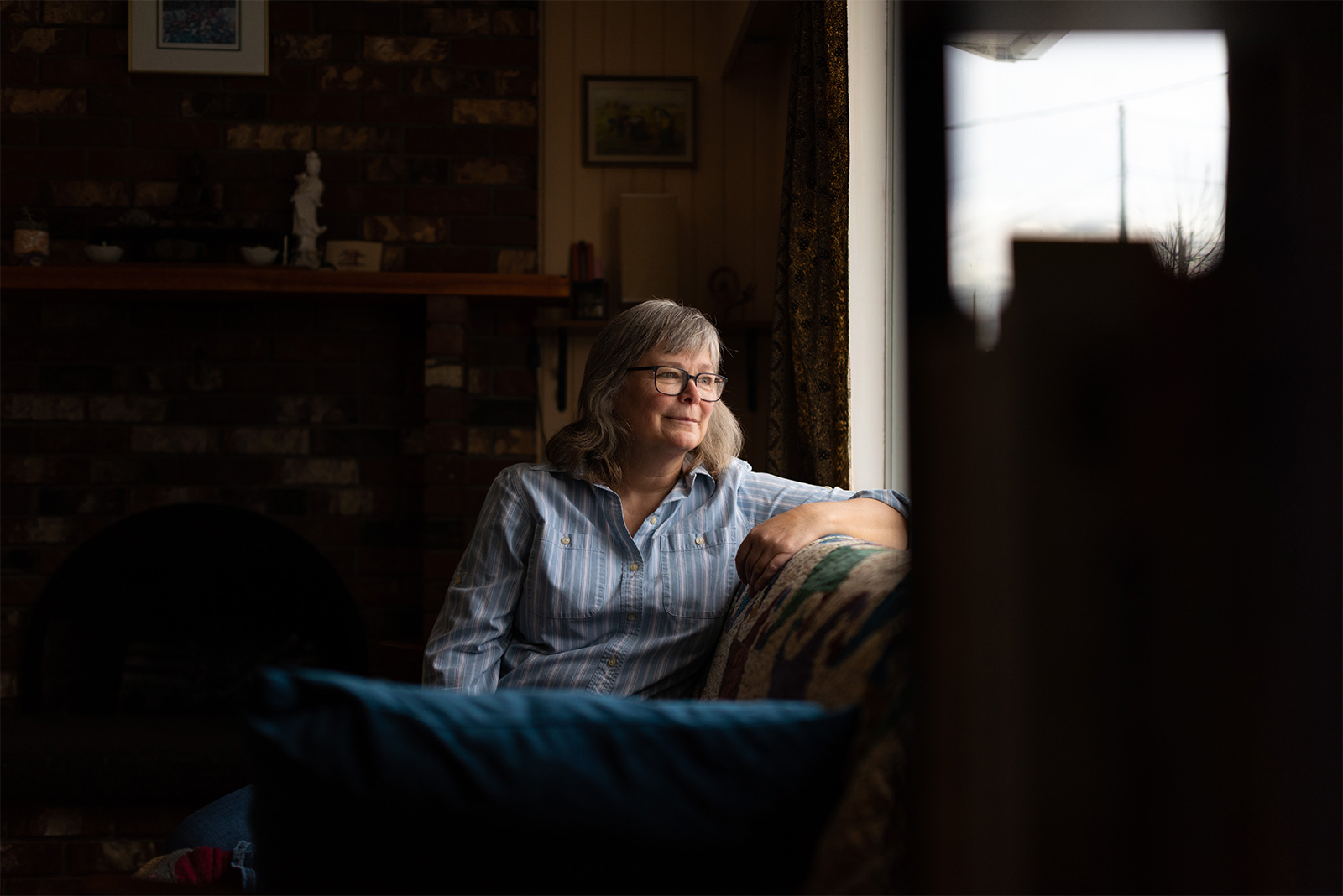 A woman sitting on a sofa in a dark room, looking out the window.