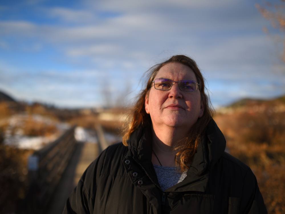 A white middle-aged woman with brown hair stand outside in a rural setting looking up. A bridge is out of focus behind her.
