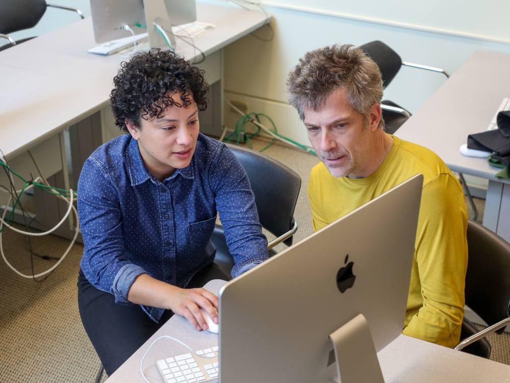 A young woman of Italian and Filipina heritage with curly dark hair sits to the left of a middle aged white man with salt and pepper hair, both looking at a computer screen with other computers around them.