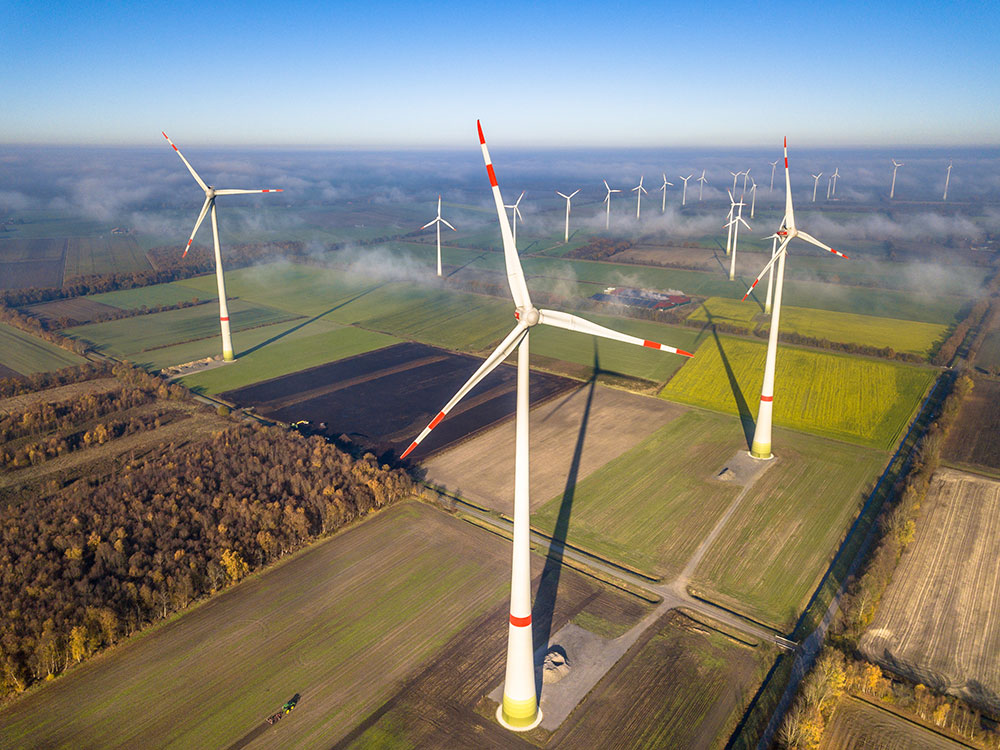 An aerial of wind tubrines amidst a piercing blue skin and farmland.