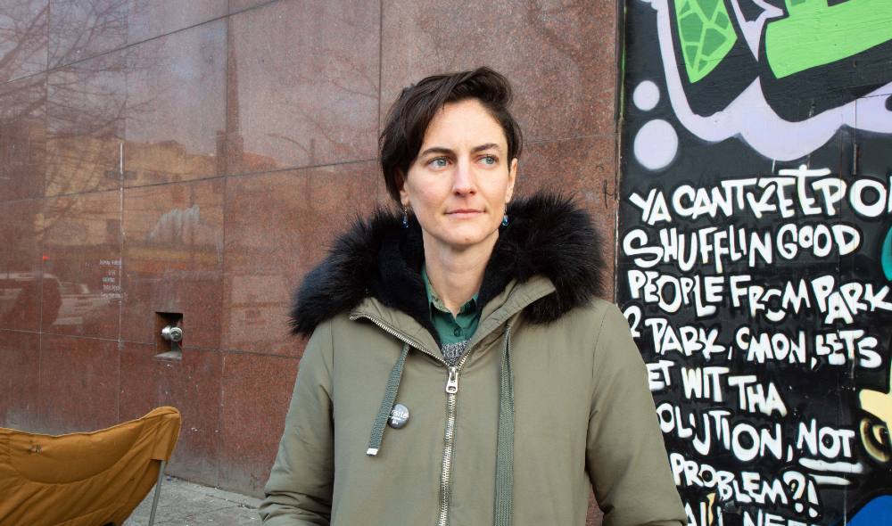 A person with short, swoopy hair, wearing a parka, earrings and a pin saying &ldquo;insite,&rdquo; stands in front of a mural.
