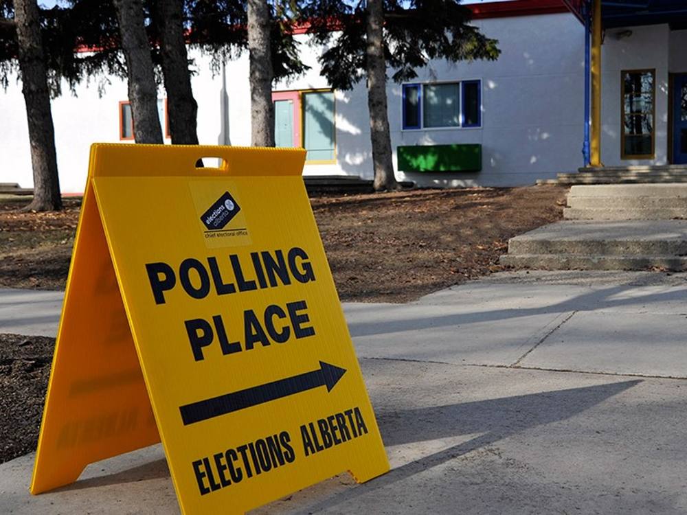 On a sunny day outside a low-rise building, a yellow sandwich board sign with and an arrow and black letters saying: Polling Place, Elections Alberta
