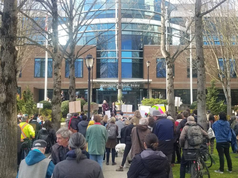 A large group of people rally outside a building on a treed lawn. Some are holding signs. At least one sign says, &ldquo;Save Ideal Mini.&rdquo;