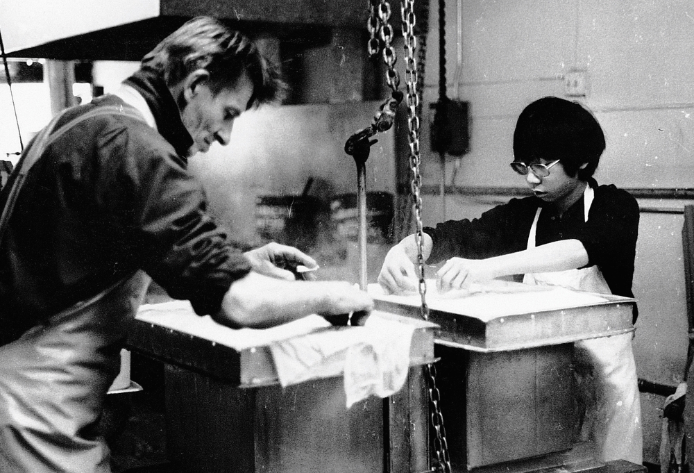 A black and white photo of a man and a young man with glasses peeling cheesecloths off of a tray of fresh tofu. They are in a steamy kitchen.