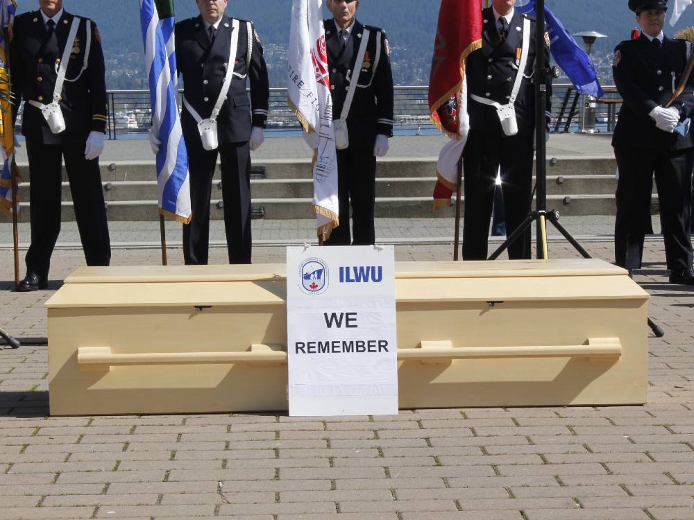 A group of people in uniform, holding flags, stand behind a plain wood coffin.