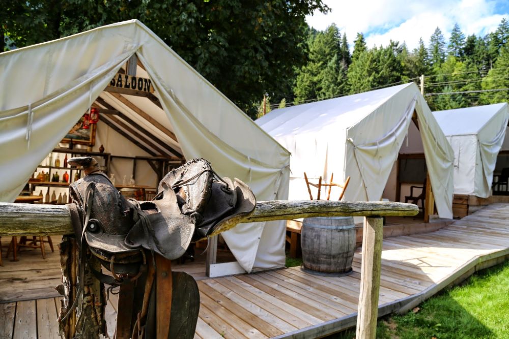 A saddle rests on an aged wooden railing in front of several tents.