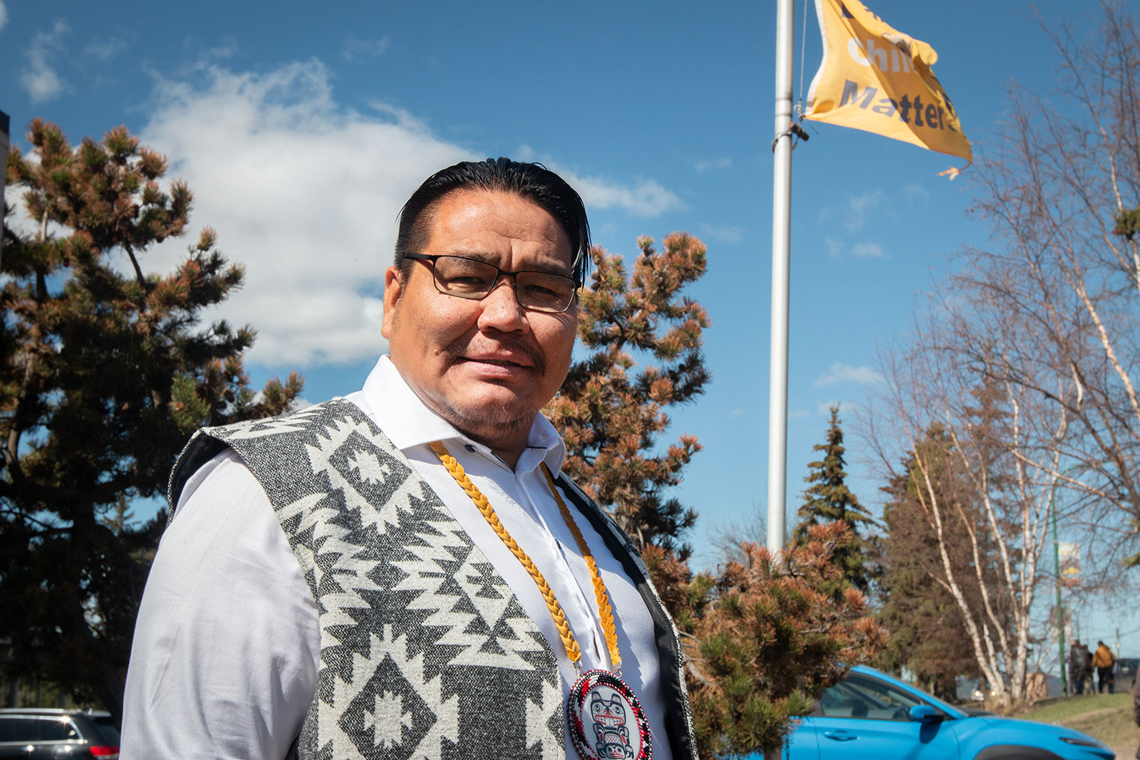 A man with dark hair and skin tone and wearing glasses stands in an outdoor setting wearing an Indigenous-style vest and pendant.