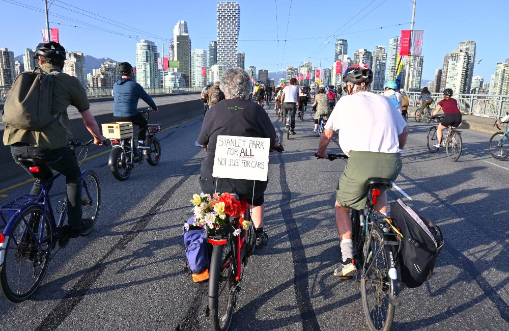 An older woman in a black t-shirt rides her bike towards downtown Vancouver with a large procession of other cyclists. She has flowers and a pannier attached to the rack of her bike. A sign also attached reads &ldquo;Stanley Park for all, not just for cars.&rdquo; 