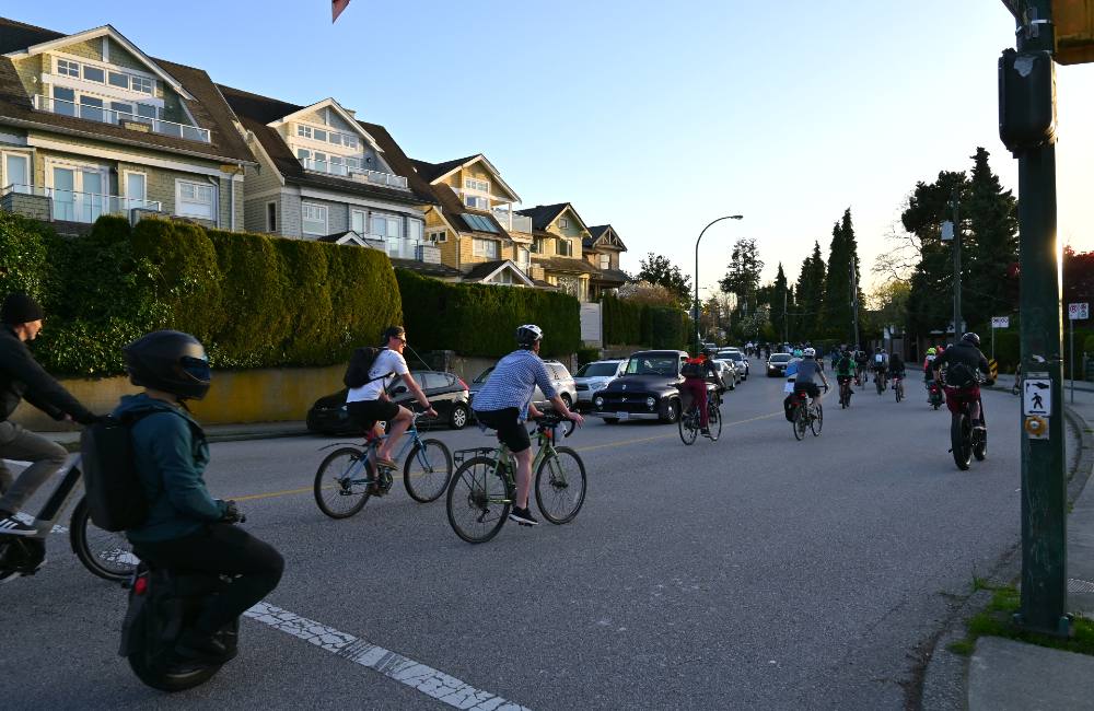 In the first image, cyclists ride on the Granville Street Bridge, many with a smile on their face. A couple of cyclists are chatting. To the left is a person on an electric scooter. Behind the group of cyclists is a line of cars. The west side of Vancouver lies behind them bordered by clear blue sky. In the second image, two cyclists chat as they ride within a procession of bikes. Behind them is a person wearing a helmet on an electric unicycle. 