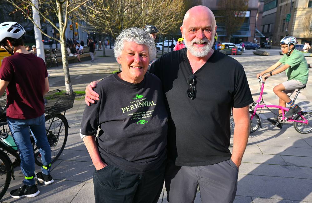 Two older people embrace at their sides surrounded by cyclists. The woman standing to the left has short grey hair and wears a black t-shirt that reads &lsquo;Not so hardy: Perennial.&rsquo; The man to the right is bald, has a grey beard and has bright blue eyes.