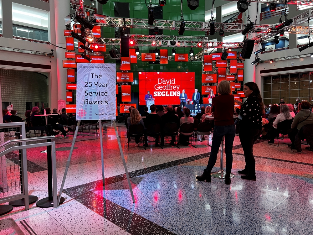 People gather in an atrium. A sign reads &lsquo;The 25 Year Service Awards.&rsquo; Screens surround a stage. On the main screen, it reads, &lsquo;David Geoffrey Seglins.&rsquo; 