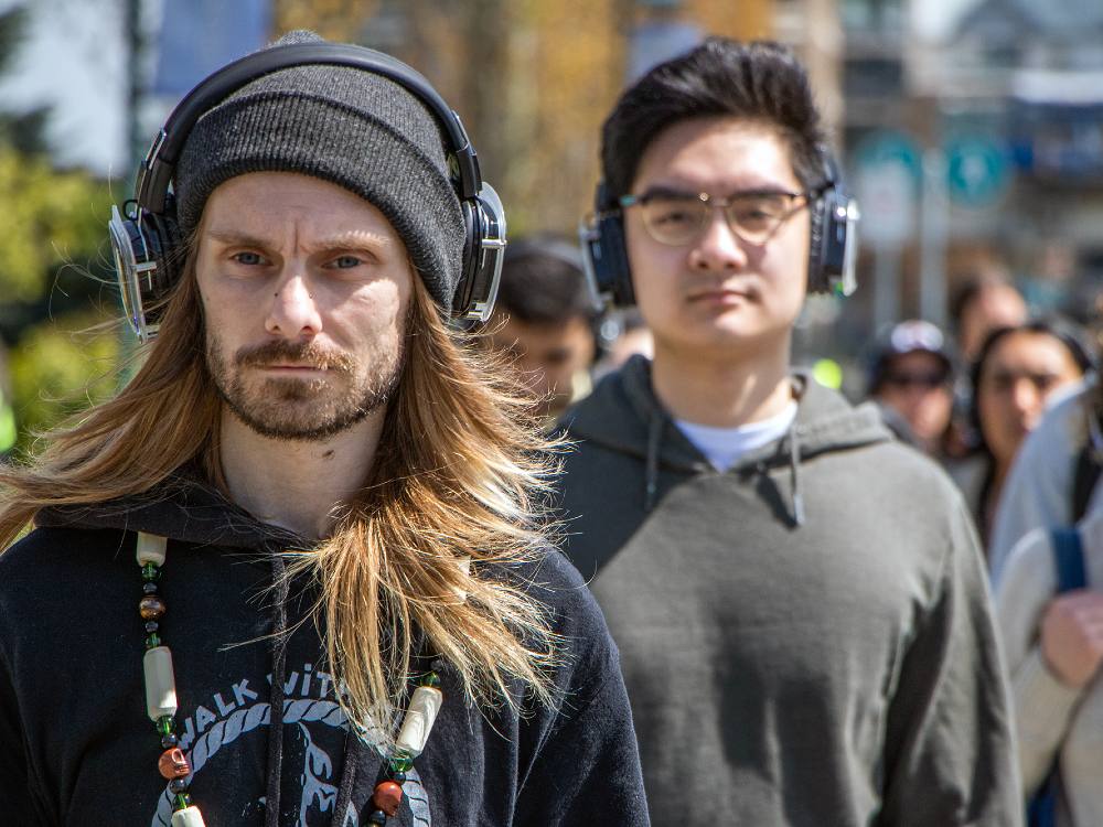 A man with long blonde hair, a black toque, headphones and a black hoodie leads a group down the False Creek seawall in Vancouver on a sunny day.