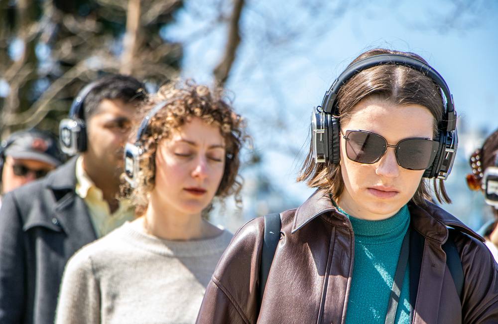 Four people participate in a &lsquo;Walk with Me&rsquo; walk in Vancouver on a sunny day. They are wearing headphones and one person with curly hair and a grey sweater is closing their eyes. Another person in front of them is wearing sunglasses and a brown leather jacket.