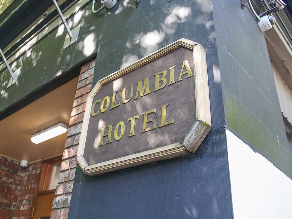 The exterior of 303 Columbia St. in Vancouver&rsquo;s Downtown Eastside features a wooden plaque bearing gold capital letters that read &ldquo;Columbia Hotel&rdquo; against a painted blue, green and white exterior. A brick alcove and doorway is visible to the left of the frame. It&rsquo;s a sunny day.