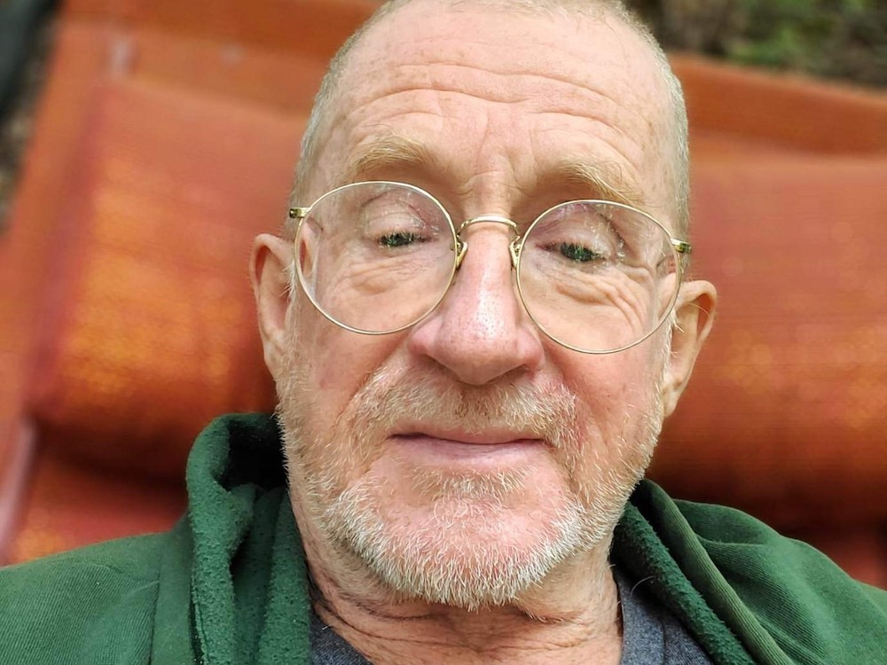 A man with a greying beard smirks into the camera. He's wearing glasses and a green jacket. 