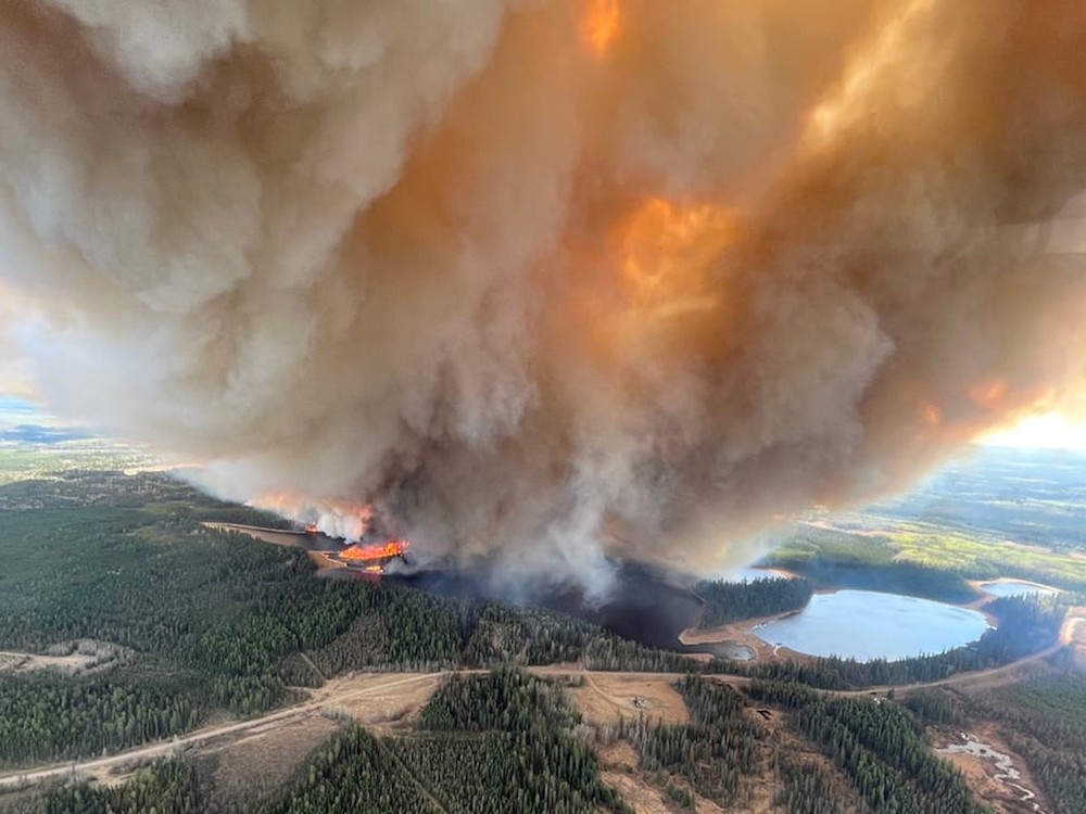 A large billowing plume of yellow-brown smoke drifts to the top of the frame next to parched earth, dense forest and a lake.