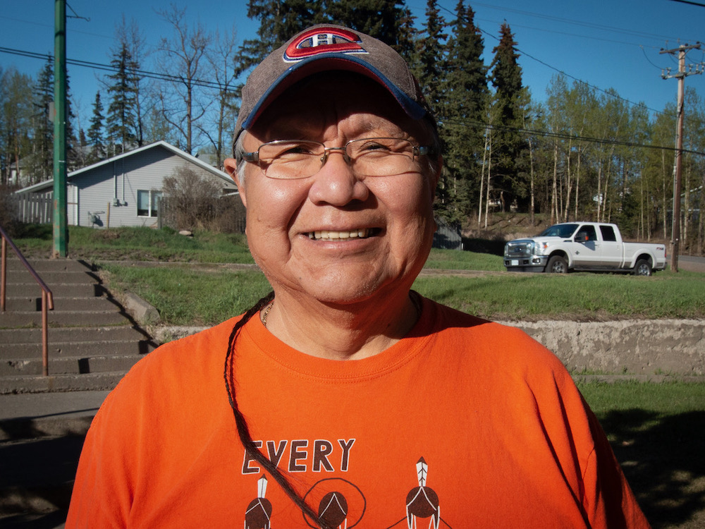 A man wearing an orange ‘Every Child Matters’ shirt and a ballcap smiles at the camera. He has a thin braid, which curves around onto his shoulder.