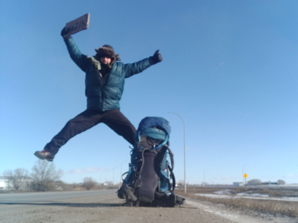 A man holding a cardboard sign by the side of the road jumps into the air, giving a thumbs up. He is wearing winter gear, the side of the road is snowy, and he&rsquo;s beside a large backpack.