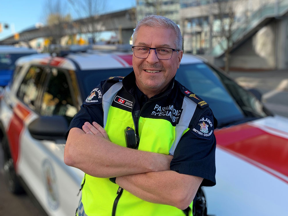 A man wearing a paramedic uniform leans against an emergency vehicle. His arms are crossed, and he smiles at the camera.