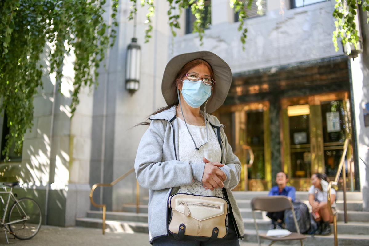 A Chinese woman in her early 60s. She&rsquo;s wearing a mask and a sun hat. She&rsquo;s standing in front of the steps of Vancouver City Hall.