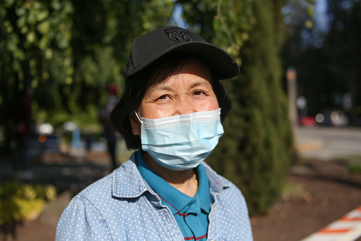 A Chinese woman in her early 60s. She&rsquo;s wearing a mask. She is wearing a white-spotted denim shirt over a teal polo.