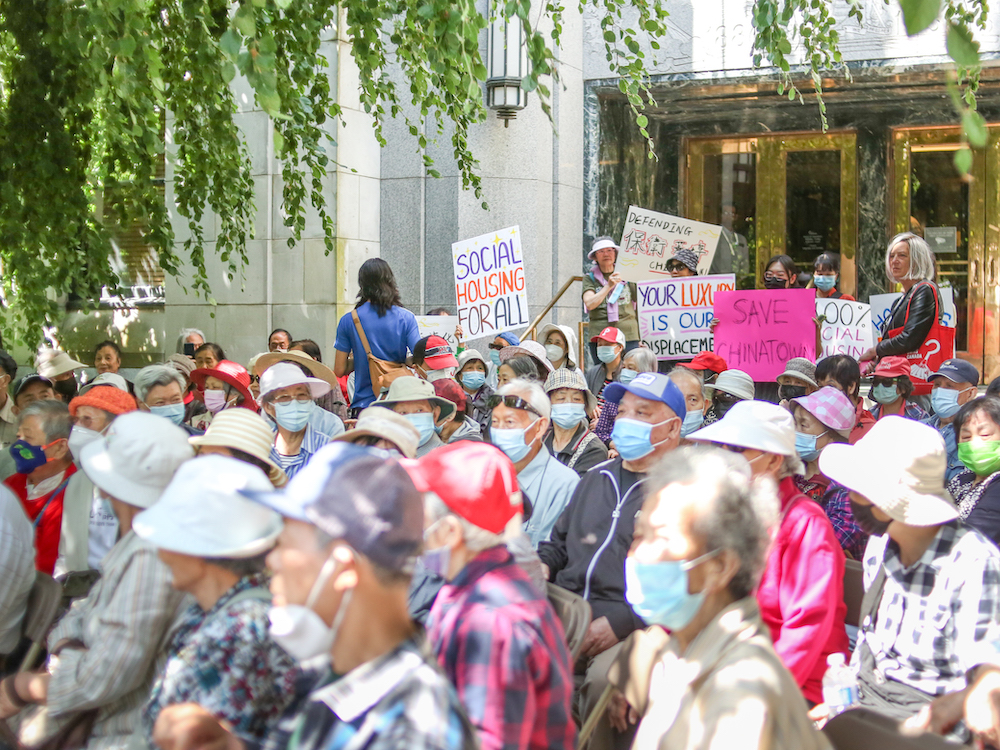 A crowd of seniors in the sun in front of Vancouver City Hall. They&rsquo;re all wearing colourful shirts and hats. There&rsquo;s a tree on the left whose branches are hanging over the crowd. The scene looks summery.